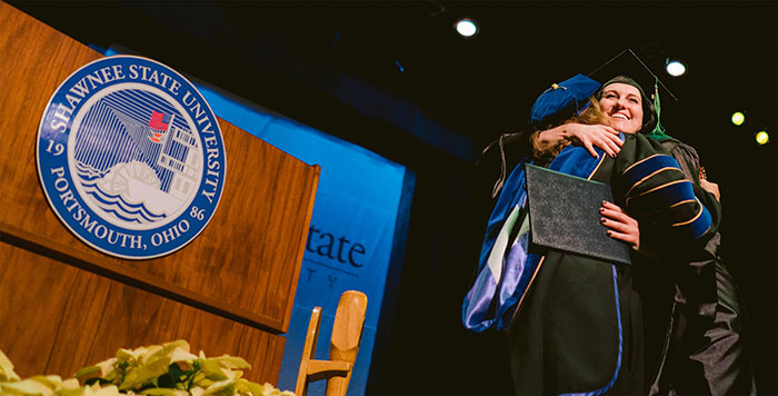 picture of student in cap and gown hugging someone on stage